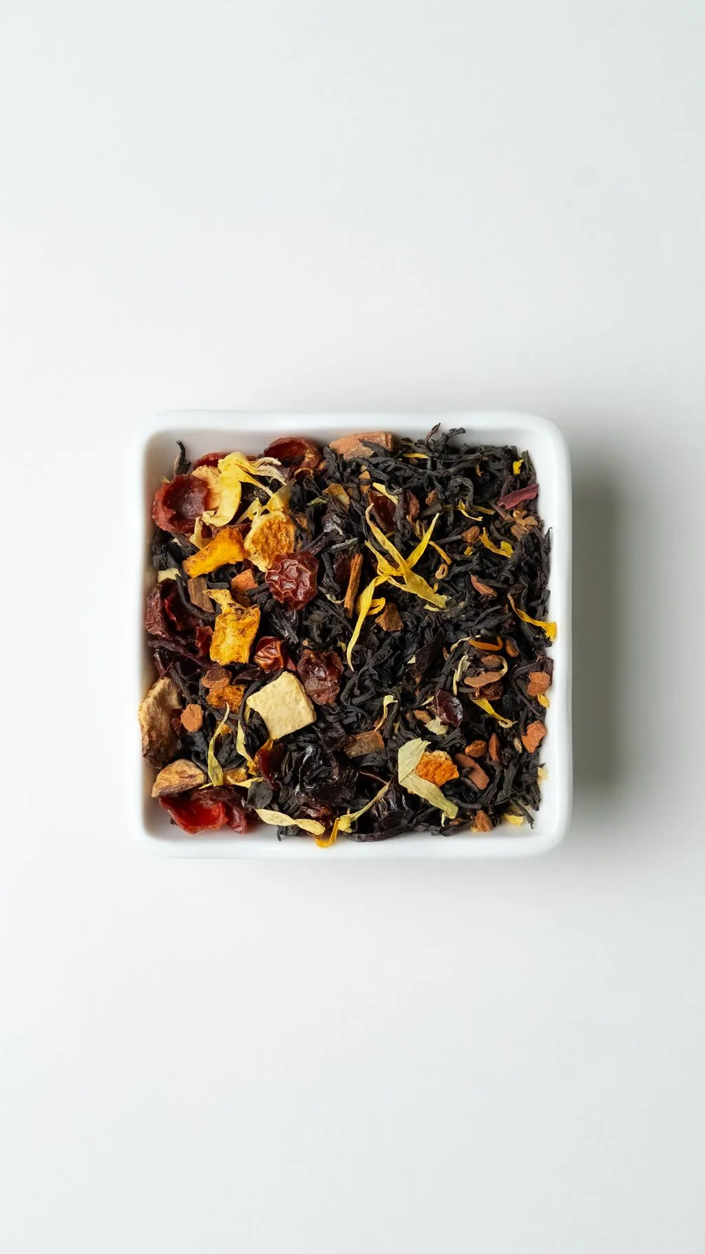Pumpkin Spice loose-leaf tea with black tea leaves, dried fruit, spices, and yellow petals in a white square dish on a clean near-white background.